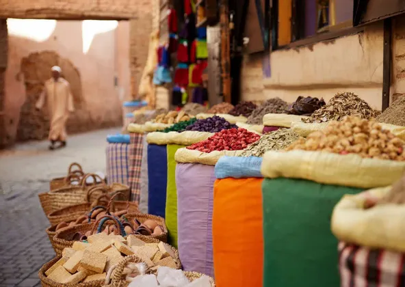 Des sacs colorés d'épices et d'herbes sont exposés à la vente sur un étal de marché extérieur dans une rue étroite.