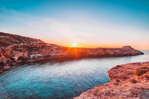 Il sole al tramonto tinge di arancione le scogliere rocciose rosse che circondano una cala tranquilla con acqua cristallina.