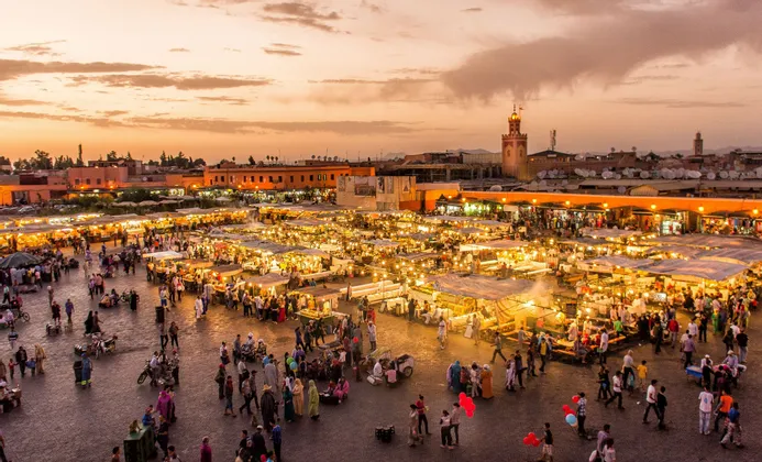 Ein erhöhter Blick auf einen belebten Marktplatz bei Sonnenuntergang, gefüllt mit Menschen, die zwischen hell erleuchteten Ständen spazieren.