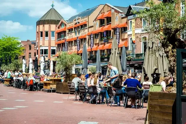 Eine belebte Straßencafé-Terrasse auf einem europäischen Stadtplatz, wo an einem sonnigen Tag Menschen an Tischen sitzen.