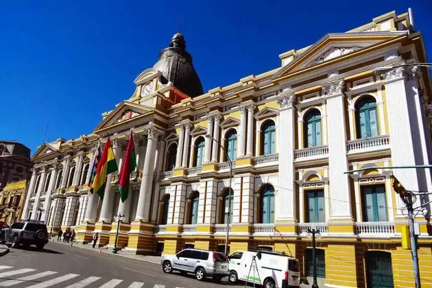 Un grande edificio neoclassico giallo e bianco con alte colonne, una cupola e diverse bandiere si erge su una strada cittadina sotto un cielo azzurro e limpido.
