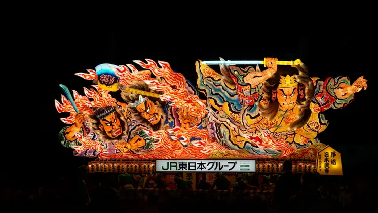 Un carro grande, colorato e illuminato che raffigura guerrieri giapponesi tradizionali con espressioni feroci a un festival notturno.