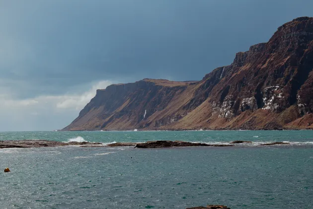 Una scogliera rocciosa e marrone con diverse cascate si affaccia su un mare turchese mosso sotto un cielo coperto.