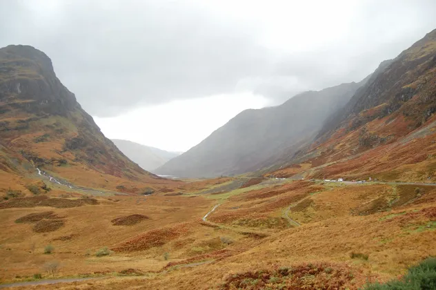 Une large vallée montagneuse avec des pentes couvertes de feuillage d'automne marron et orange sous un ciel couvert.