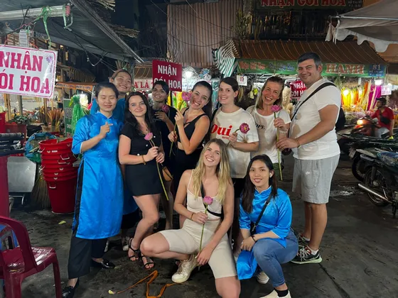 Eine WeRoad-Gruppenreise posiert für ein Foto auf einem Nachtmarkt, wobei viele Mitglieder rosa Lotusblumen halten.
