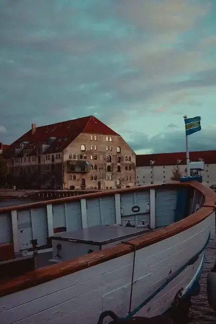 Une vue depuis un bateau en bois montre de grands et vieux bâtiments en briques le long d'un quai sous un ciel nuageux au crépuscule.
