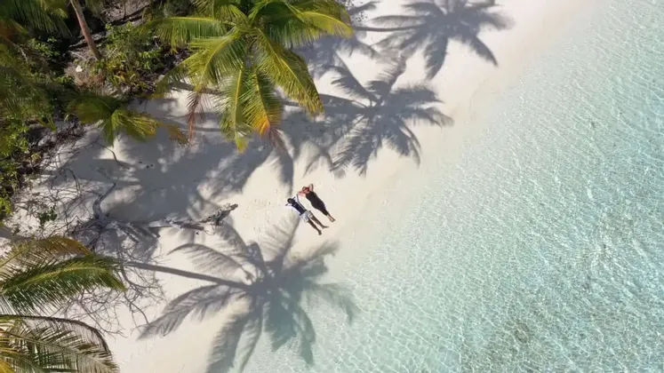 Una vista aerea di due persone che si rilassano su una spiaggia di sabbia bianca, con acqua cristallina e ombre di palme.