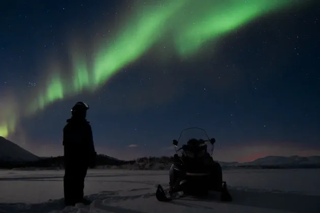 Una persona si trova accanto a una motoslitta in un paesaggio innevato di notte, osservando l'aurora boreale verde riempire il cielo stellato.