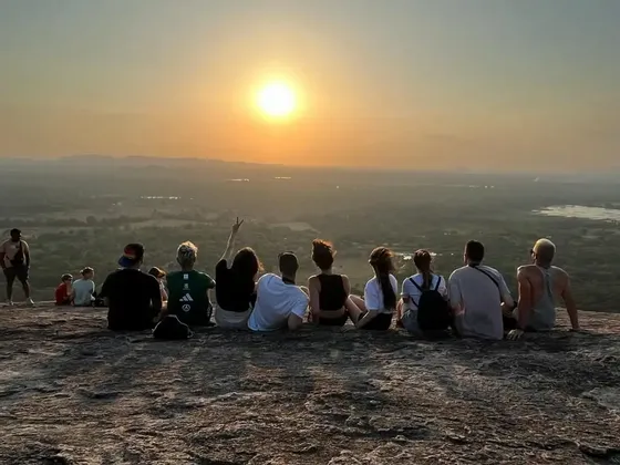 Un grupo de WeRoad, sentado en un promontorio rocoso, contempla la puesta de sol sobre un extenso valle verde y colinas distantes.