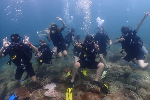 Un gruppo WeRoad in attrezzatura subacquea posa per una foto sottomarina, facendo segni con le mani sopra una scogliera rocciosa.