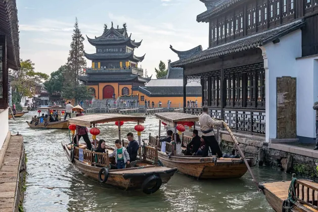 Persone su barche di legno percorrono un canale fiancheggiando edifici tradizionali e una grande pagoda.
