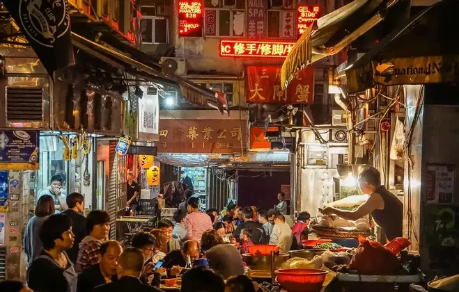Una grande folla di persone che mangiano ai tavoli all'aperto in un affollato vicolo cittadino di notte, illuminato da insegne al neon e bancarelle di cibo.