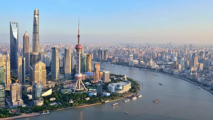 Una vista aerea dello skyline di una città moderna con numerosi grattacieli, inclusa una torre prominente, che costeggiano un ampio fiume sotto un cielo nebbioso.