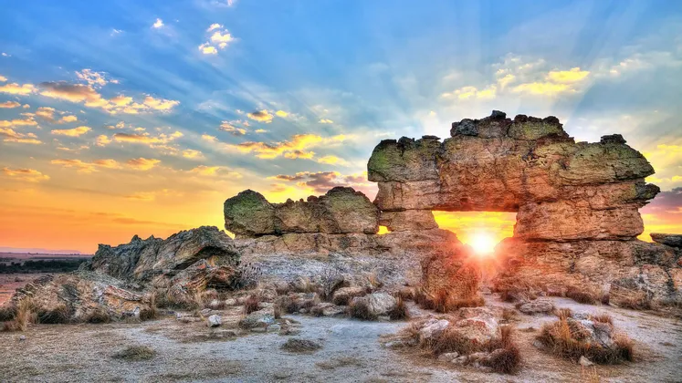 Il sole tramonta dietro una grande formazione rocciosa, la sua luce splende luminosa attraverso un arco naturale sotto un cielo colorato.