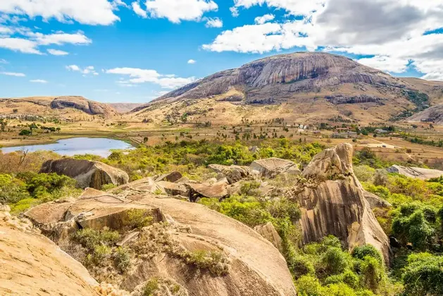 Un'ampia valle con un lago e grandi massi, che conduce a un'imponente montagna rocciosa sotto un cielo blu parzialmente nuvoloso.