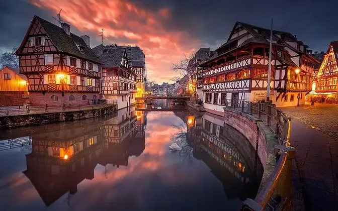 Beleuchtete Fachwerkhäuser säumen einen Kanal, deren Spiegelungen und ein farbenprächtiger Sonnenuntergang sich im Wasser widerspiegeln.