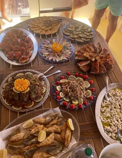 Un tavolo di legno ospita numerosi vassoi di cibo, tra cui pesce fritto, bruschette, verdure grigliate e varie insalate al limone.