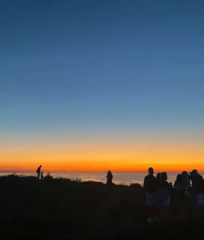Sagome di un viaggio di gruppo WeRoad su una collina che osservano il tramonto arancione e blu sul mare.
