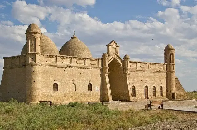 Un edificio storico color sabbia con cupole multiple e un grande ingresso ad arco si trova in un paesaggio erboso sotto un cielo nuvoloso