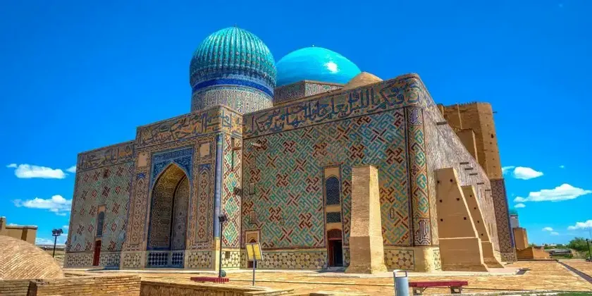 Un mausoleo sfarzoso, con due cupole blu e pareti ricoperte da intricati mosaici di piastrelle blu e verdi, si erge sotto un cielo azzurro limpido.