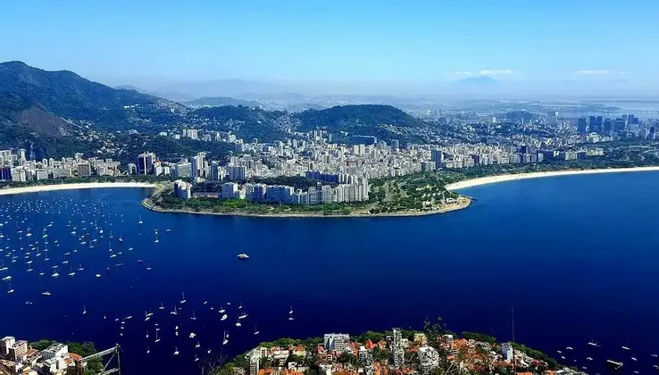 Vue aérienne d'une ville côtière le long d'une baie remplie de voiliers, nichée contre des collines verdoyantes sous un ciel bleu clair.