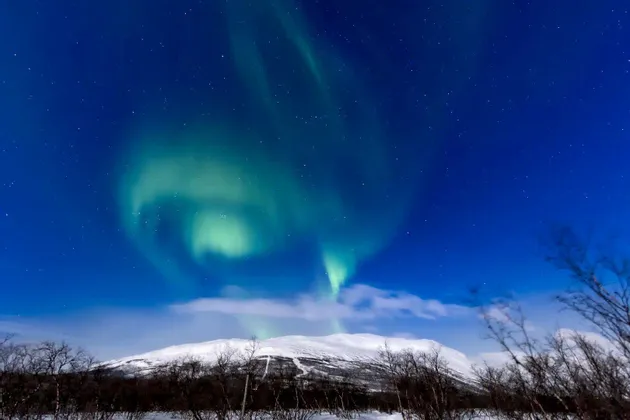 La aurora boreal verde se arremolina en un cielo nocturno estrellado sobre un paisaje montañoso cubierto de nieve.