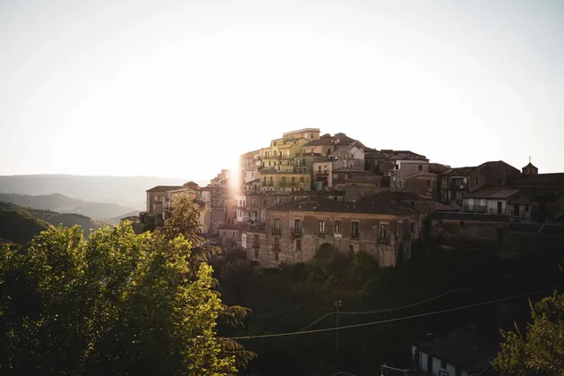 Ein Dorf mit alten Häusern, erbaut an einem grünen Hang, wobei die aufgehende Sonne einen hellen Schein hinter den Gebäuden erzeugt.