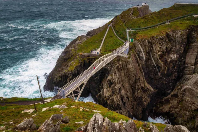 Eine moderne Fußgängerbrücke überquert eine Schlucht zwischen zwei steilen, grasbewachsenen Klippen, während Menschen darüber über einem stürmischen Meer gehen.