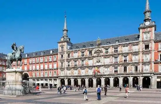 Una statua equestre si erge in un'ampia piazza pavimentata, di fronte a un elaborato edificio storico con guglie, sotto un cielo azzurro e limpido.