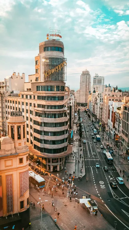 Vista aerea di una trafficata via cittadina con traffico, pedoni e un prominente edificio curvo con grandi insegne sotto un cielo nuvoloso.