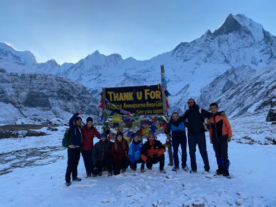 Un viaje en grupo de WeRoad posa para una foto frente al letrero del Campamento Base Annapurna, rodeado de montañas cubiertas de nieve.