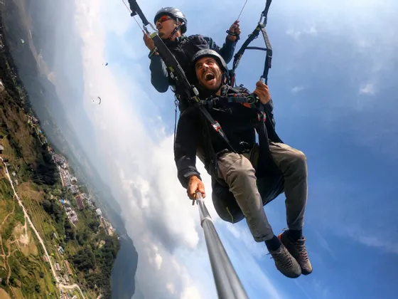 Un hombre se toma una selfie mientras hace parapente en tándem muy por encima de un valle verde y montañoso.