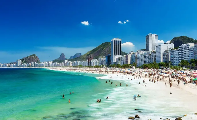 Une plage urbaine animée avec des gens nageant dans l'eau turquoise et se relaxant sur le sable blanc, avec des montagnes en arrière-plan.