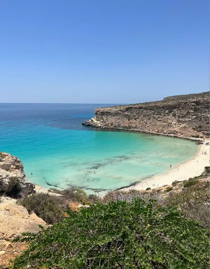 Una vista dall'alto di una cala appartata con acqua turchese e una spiaggia di sabbia bianca, circondata da scogliere rocciose sotto un cielo sereno.