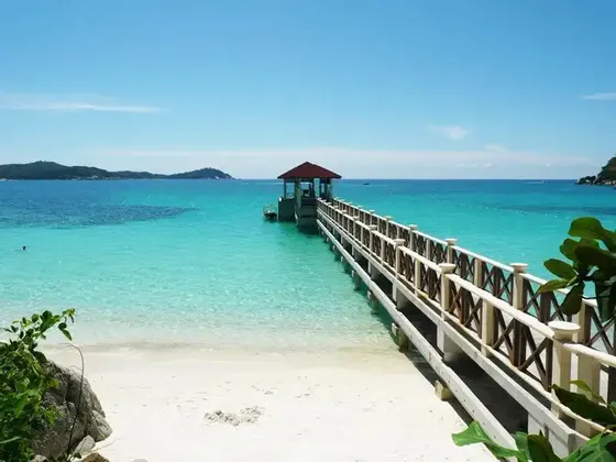 Un pontile di legno si estende da una spiaggia di sabbia bianca in acque cristalline turchesi, con isole lontane sotto un cielo azzurro brillante.