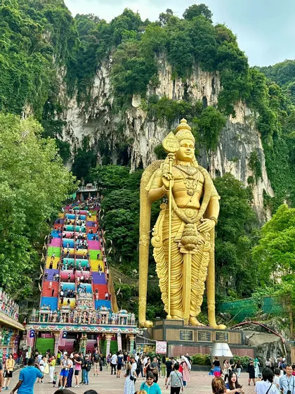 Una gigantesca statua dorata di una divinità si erge accanto a una lunga e colorata scalinata dove le persone salgono su una montagna lussureggiante e verde.