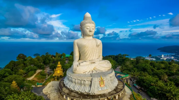 Eine große, weiße Stein-Buddha-Statue sitzt auf einem grünen Hügel mit Meerblick unter einem strahlend blauen Himmel.