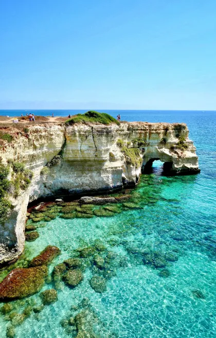 Una vista costiera con scogliere bianche e un arco di roccia naturale su acque limpide color turchese in una giornata di sole.