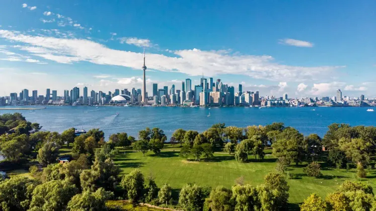 Uno skyline cittadino con numerosi grattacieli al di là di uno specchio d'acqua, osservato da un rigoglioso parco verde in una giornata di sole.