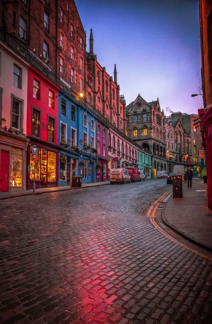 Una calle adoquinada curva bordeada de edificios coloridos y tiendas al anochecer, con una luz roja reflejándose en las piedras.