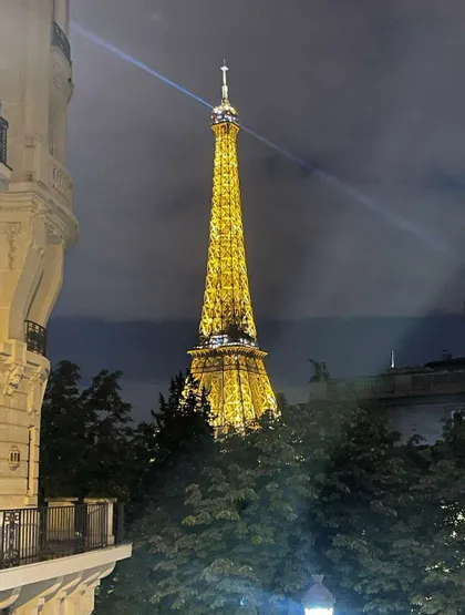 La Torre Eiffel illuminata da luci dorate di notte, il suo faro che proietta un fascio nel cielo nuvoloso, visibile sopra le cime degli alberi.