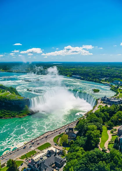 Una veduta aerea di una vasta cascata a ferro di cavallo che si riversa in un fiume turchese, con una strada e una città immerse nel lussureggiante paesaggio verde.