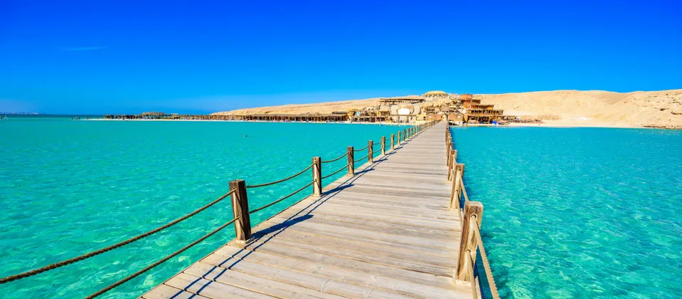 Un lungo pontile in legno con ringhiere di corda si estende su acque limpide color turchese verso una spiaggia sabbiosa con edifici sotto un cielo blu.
