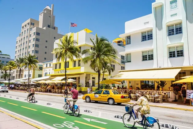 Le persone vanno in bicicletta su una pista ciclabile verde lungo una strada soleggiata fiancheggiata da edifici Art Déco e palme.