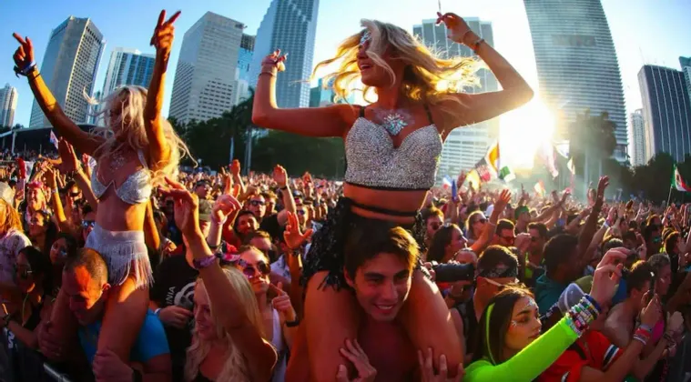 Una grande folla di persone balla a un festival musicale all'aperto, con donne sulle spalle, sullo sfondo di uno skyline cittadino.