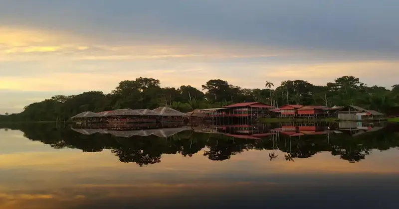 Un villaggio di palafitte su un lago sereno riflette perfettamente la giungla circostante e un cielo al tramonto colorato.