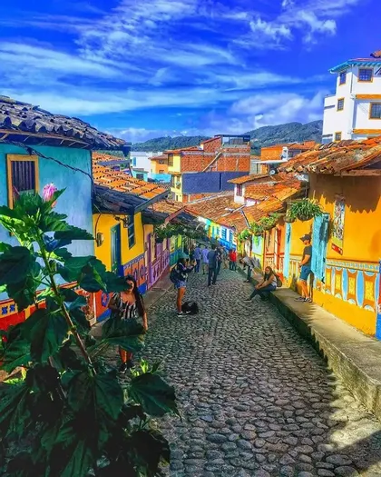 Una veduta dall'alto di una strada acciottolata in pendenza, fiancheggiata da edifici dai colori vivaci, con persone che passeggiano sotto un cielo azzurro.
