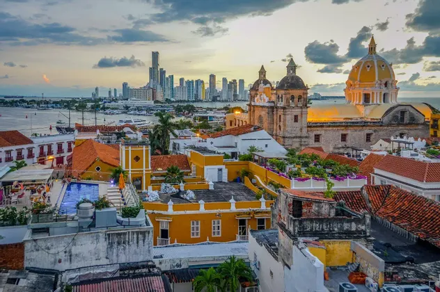 Veduta urbana di edifici storici con tetti in terracotta e una cattedrale, con uno skyline moderno sull'acqua al tramonto.