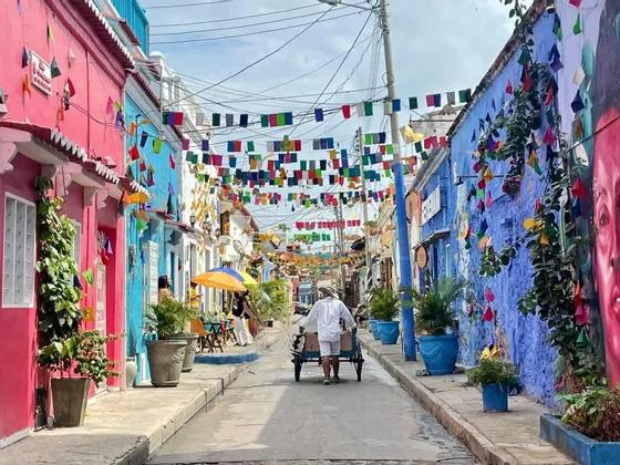Un uomo spinge un carretto lungo una strada stretta fiancheggiata da edifici colorati e bandiere festive appese in alto.