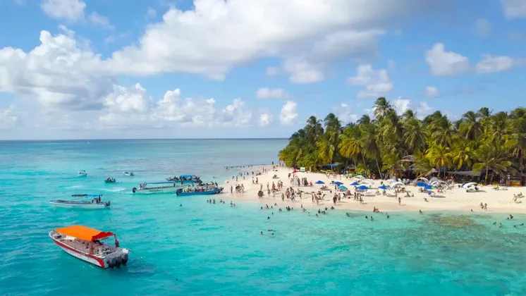 Una vista aerea di una spiaggia tropicale affollata con acqua turchese, numerose imbarcazioni e persone che nuotano e prendono il sole sulla sabbia.